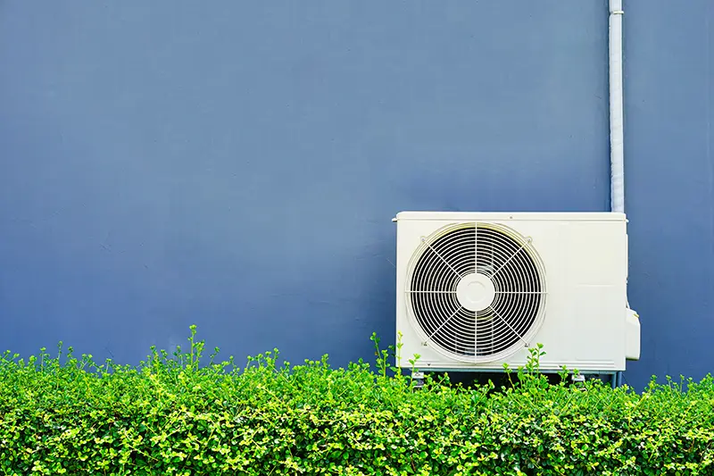 White outdoor air conditioning unit against a dark blue wall with a lush green hedge in front.